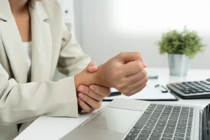 Foto em close do antebraço de uma mulher segurando o próprio pulso com a outra mão, em frente ao computador, demonstrando dor na articulação.