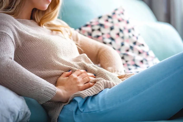 Mulher sentada no sofá, mãos no abdômen, expressando dor.