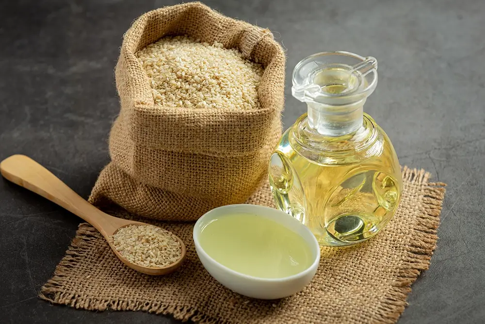 Foto artística de sementes de gergelim em um delicado e pequeno saco ao lado de um frasco com óleo de gergelim, uma colher com sementes de gergelim e uma combuca com molho, Todos os elementos estão sobre um pano de juta, produzindo uma atmosfera de muito bom gosto.