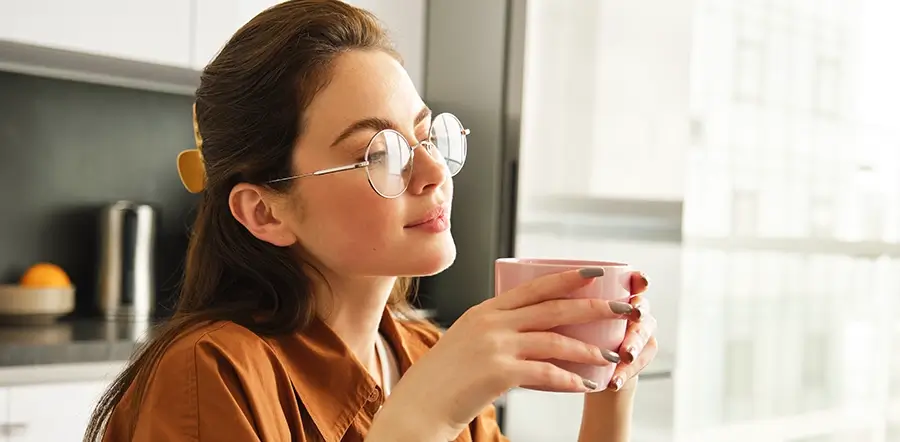 Bonita jovem segura uma xícara de café enquanto parece estar contemplando a paisagem pela janela de sua casa.