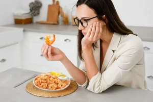 Mulher observando um camarão antes de comer. Ela está diante de um prato com camarões. A imagem ilustra uma dúvida comum de quem tem alergia a camarão