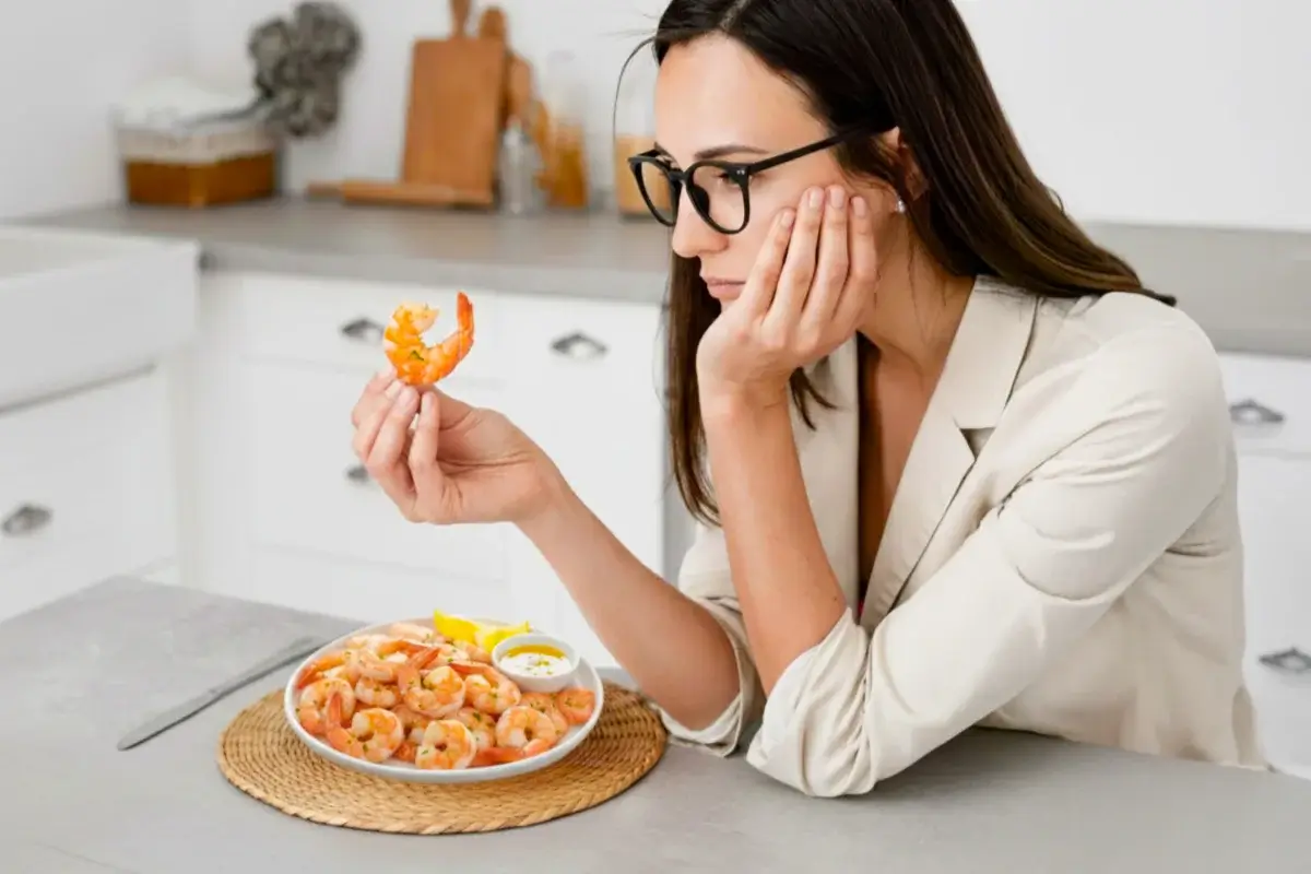 Mulher observando um camarão antes de comer. Ela está diante de um prato com camarões. A imagem ilustra uma dúvida comum de quem tem alergia a camarão